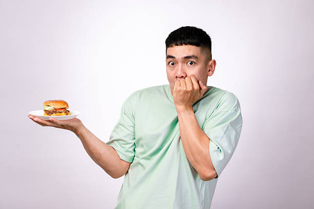 istock An Asian man in a mint green shirt holds a plate with a cheeseburger while making a disgusted expression, sticking out his tongue and touching his neck. The background is plain white 2203437348