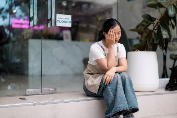 istock Weary Asian server resting head on hand, displaying stress near shuttered cafe during challenging economic period 2203437576