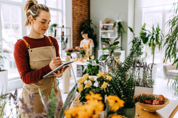 istock Young florist working in a vibrant flower shop while taking notes and creating beautiful arrangements with a coworker nearby 2203439998