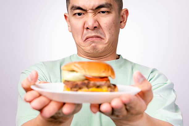 istock An Asian man in a mint green shirt holds a plate with a cheeseburger while making a disgusted expression. The background is white 2203441515