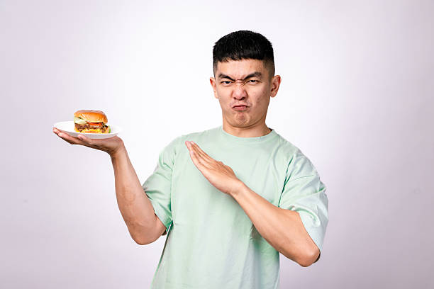 istock An Asian man in a mint green shirt holds a plate with a cheeseburger while extending his other hand in a stop gesture. He looks surprised or disapproving. The background is plain white 2203442007