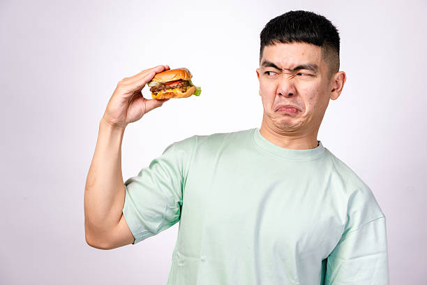 istock An Asian man in a mint green shirt holds a plate with a cheeseburger while extending his other hand in a stop gesture. He looks surprised or disapproving. The background is plain white 2203442869