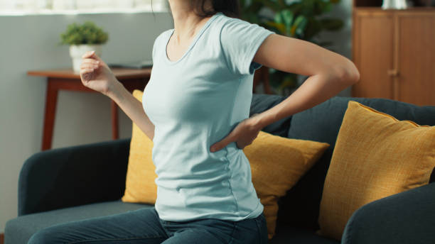 istock Asian woman hand holding her low back, feeling low back pain and hurt while sits on sofa in living room at home 2203447916