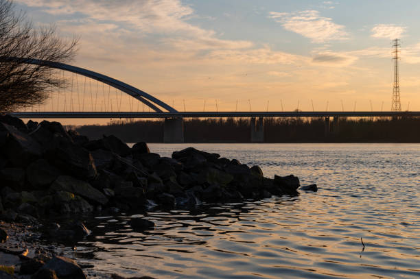 A peaceful sunset scene featuring the Pentele Bridge crossing the Danube River. The warm light reflects on the water, while dark rocks in the foreground add contrast to the tranquil landscape.