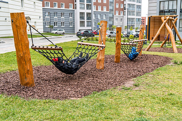 Kids lying on black net hammocks in a modern residential playground with wooden structures, surrounded by green grass and apartment buildings