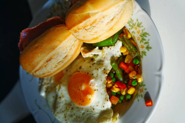 A sunny-side-up egg rests beside a vibrant vegetable medley, crispy bacon, and two golden rolls on a floral-rimmed plate, capturing a simple, hearty breakfast or brunch.