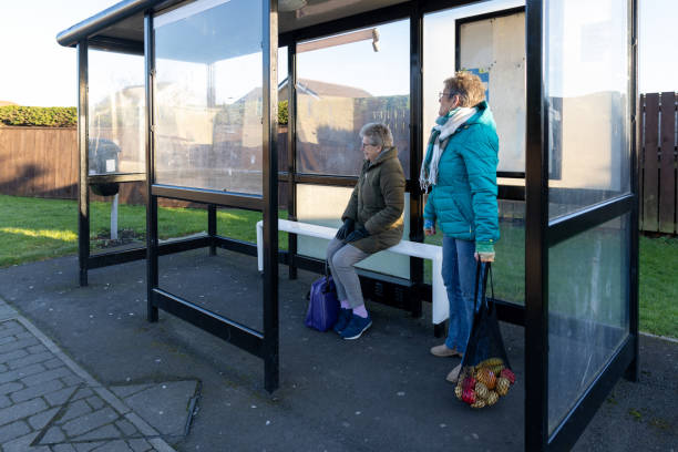 Wide shot of two senior female friends waiting inside a bus shelter in Newcastle-upon-Tyne, North East England. They are both wearing warm winter coats and eyeglasses, looking off-frame as they wait for a bus, one holding a net bag filled with fruit. One of the females is living with dementia.\n\nVideos similar to this scenario available.