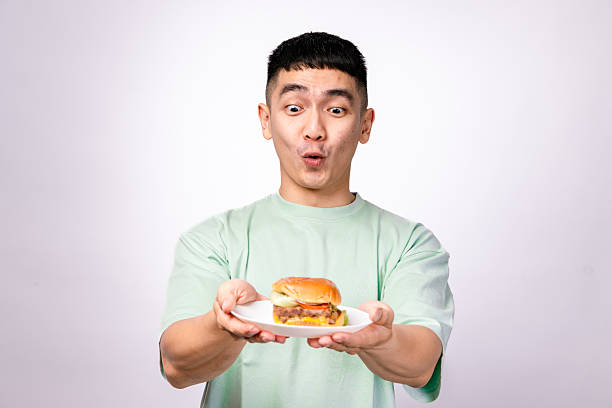 An Asian man in a mint green shirt excitedly presents a cheeseburger on a white plate. He has a wide smile, and the background is plain white