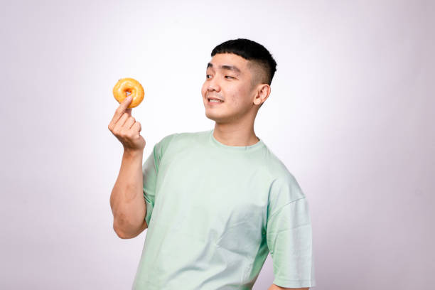 istock An Asian man in a mint green shirt holds a glazed donut with a curious expression, looking at it against a white background, symbolizing enjoyment, food cravings, or a love for sweets 2203464210