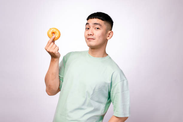 istock An Asian man in a mint green shirt holds a glazed donut with a curious expression, looking at it against a white background, symbolizing enjoyment, food cravings, or a love for sweets 2203464236