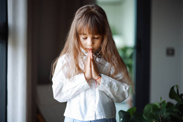 A child girl closes her eyes and clasps her hands together in prayer, expressing her faith and seeking solace in a moment of quiet reflection and spiritual connection.