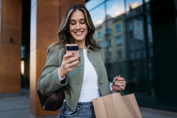istock Smiling young woman using smartphone while carrying shopping bag 2203478399