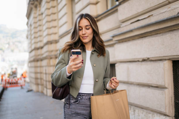 Young stylish woman using smartphone while carrying shopping bag