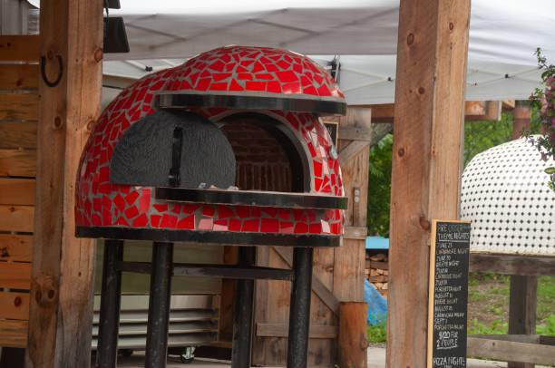 A vibrant red mosaic-covered wood-fired pizza oven is located outdoors, blending with natural wooden surroundings. Designed to prepare artisanal pizzas, emphasizing culinary art and rustic aesthetics in an outdoor ambiance.