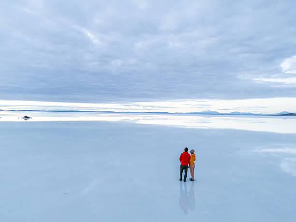 Couple stands together on reflective salt flats. Embracing duo surrounded by endless horizon. Dreamlike landscape with vast open sky.