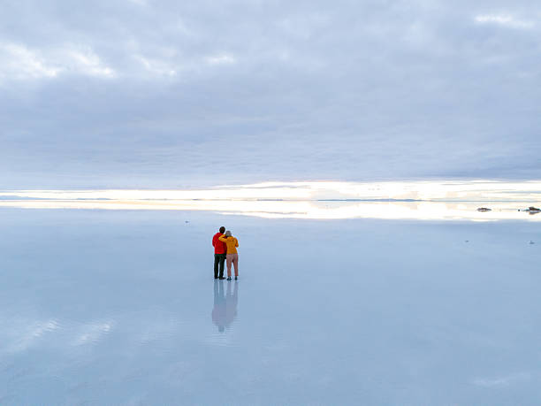 Couple stands together on reflective salt flats. Embracing duo surrounded by endless horizon. Dreamlike landscape with vast open sky.