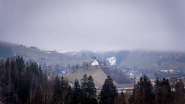 A misty valley with a small church on a hill, surrounded by trees, distant mountains, and a foggy atmosphere.