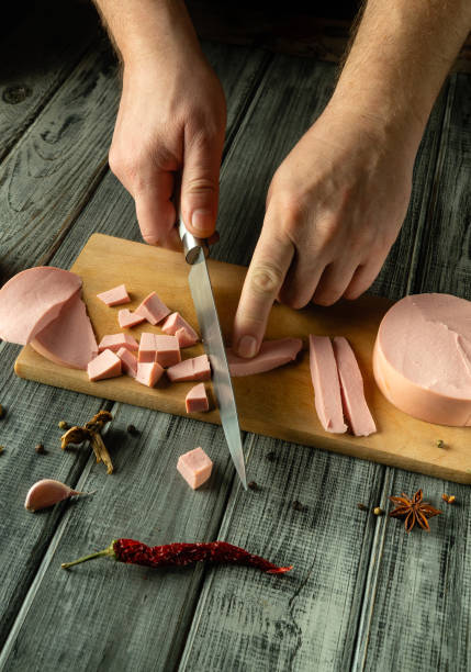 istock Hands expertly slice a slab of sausage for lunch, creating neat cubes and strips on a rustic wooden cutting board surrounded by vibrant spices and chili peppers 2203496196