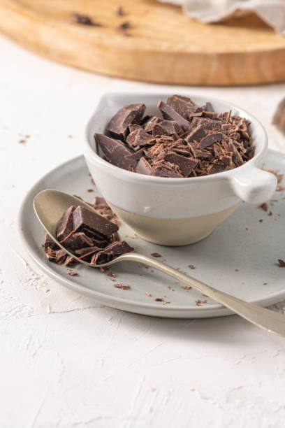 istock Chocolate chunks in a white ceramic cup on a matching saucer with a spoon, set on a textured white surface. The background includes a wooden board 2203525378