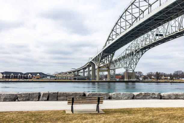 A scenic view featuring pedestrians along the river under the Bluewater Bridge in Sarnia, Ontario. The bridge stretches magnificently across the water, connecting scenic landscapes.