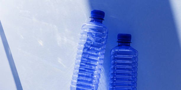 Transparent plastic bottles with blue caps and sun shadows on light background. Top view banner with Water bottles at natural light. Themes of recycling, sustainability, water conservation