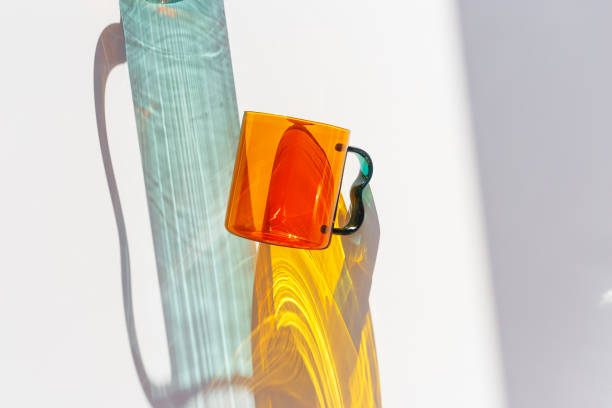 istock Orange Glass Mug with bright sunlight Shadows, transparent mug casting dynamic, vibrant lights reflections and shadows on light white surface, creative aesthetic top view, minimal close up 2203534053