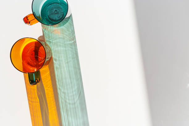 Orange and green Glass Mugs with bright sunlight Shadows, transparent cups casting dynamic, vibrant lights reflections and shadows on light white, creative aesthetic top view, minimal style, closeup