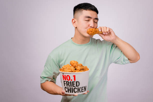istock An Asian man in a light green shirt enjoys a crispy fried chicken drumstick while holding a large bucket labeled No.1 Fried Chicken. He looks happy and satisfied. The background is white 2203537415