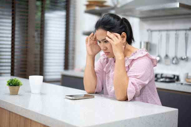 istock Woman in kitchen with stressed expression sitting at table holding head next to a phone and coffee cup 2203553504