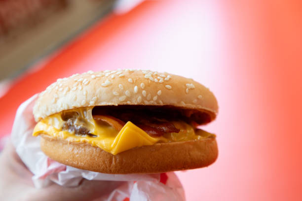 A person is holding a hamburger with cheese and bacon. The hamburger is on a bun and is being held in a paper wrapper