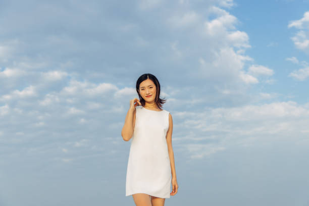 A woman standing confidently outdoors against a backdrop of a cloudy sky. She wears a white dress, exuding elegance and simplicity, while emanating a calm and serene demeanor. The image captures the beauty of nature as well as human grace and confidence in a harmonious setting.