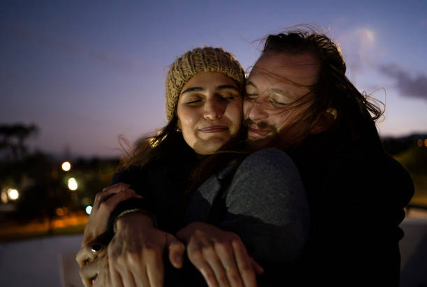 Close-up of romantic young couple standing together in a park at night, with the man hugging the woman from behind