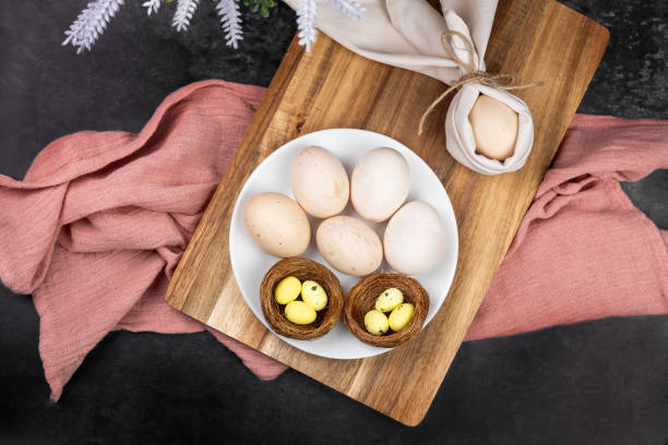 Natural farm fresh eggs on a plate on a dark background, next to decorative painted quail eggs in a nest, flowers, preparation for Easter, painting eggs for Easter, farm concept