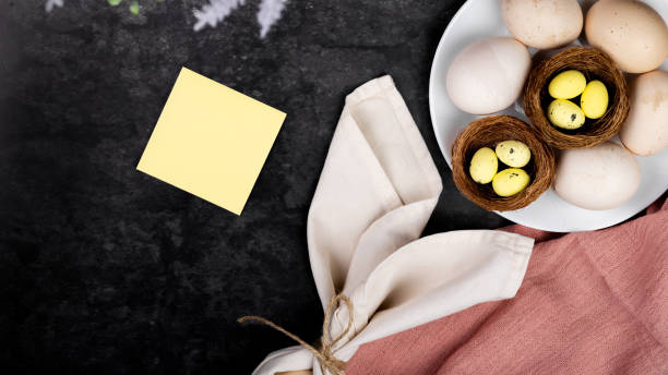 istock Natural farm fresh eggs on a plate on a dark background, next to decorative painted quail eggs in a nest, flower. Place for text - sheet of paper, copy space. Preparation for Easter 2203575733