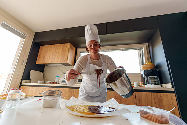 Woman pastry chef pouring melted chocolate cream on a cake base using a blender, in a modern kitchen