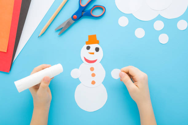 istock Little child boy hands creating and gluing snowman shape from white paper on light blue table background. Pastel color. Making winter elements. Closeup. Point of view shot. Top down view. 2203588419