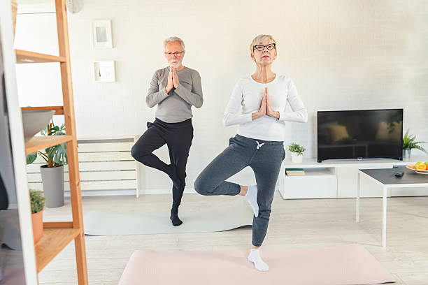 istock Senior couple practicing yoga at home, standing in tree pose with hands in prayer position. They are balancing on one leg in a bright, modern living room 2203589244