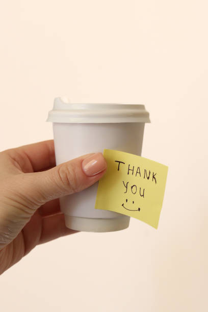 istock A person's hand holding a white paper cup with a plastic lid. A yellow sticky note with the inscription Thank you and a smiling emoticon is attached to the cup. The background is light. 2203594035