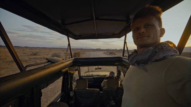 Caucasian male safari professional documents adventure in khaki attire using smartphone while steering safari vehicle through Kenya's rugged terrain during golden hour.