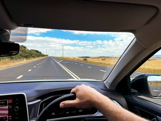 istock A wide-angle view through a car’s windscreen reveals a highway through a desert plain, framed by the interior, steering wheel, and driver’s hands under a clear blue sky with fluffy clouds 2203605656