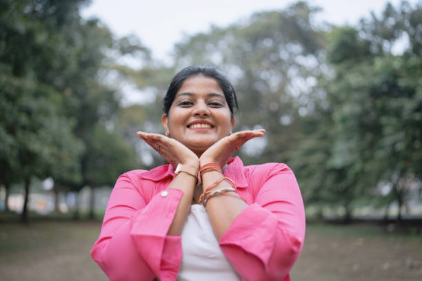 A cheerful young woman wearing a pink shirt and white top poses playfully with her hands framing her face while smiling brightly. She stands in a lush green park with trees in the background, exuding joy and confidence. The natural outdoor setting adds a fresh and vibrant feel to the image. Ideal for concepts related to happiness, positivity, self-confidence, and outdoor leisure.