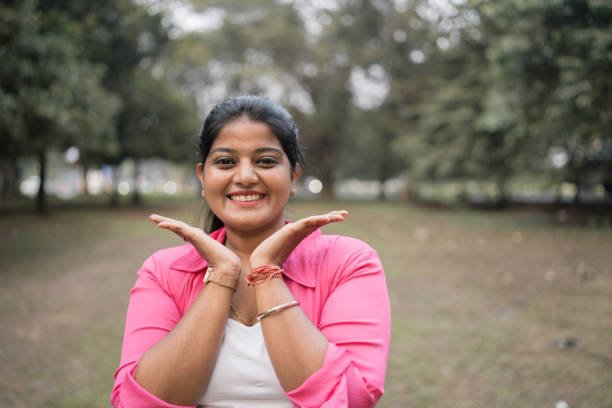 A cheerful young woman wearing a pink shirt and white top poses playfully with her hands framing her face while smiling brightly. She stands in a lush green park with trees in the background, exuding joy and confidence. The natural outdoor setting adds a fresh and vibrant feel to the image. Ideal for concepts related to happiness, positivity, self-confidence, and outdoor leisure