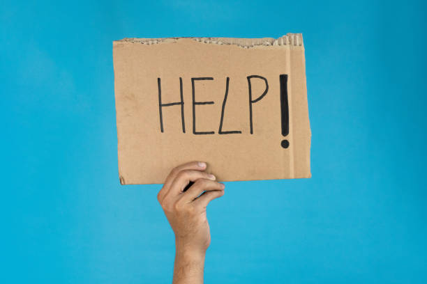 A hand holds a cardboard sign with HELP! written on it against a blue background, signifying a plea for assistance and support.