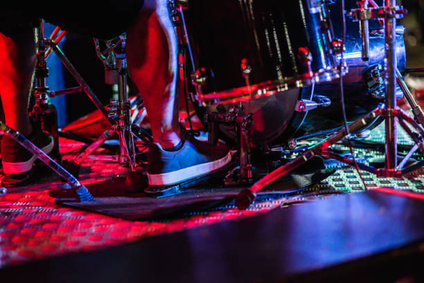 Close-up shot of a drummer's feet pressing the bass drum pedal during a live performance. Colored stage lighting reflecting on the drum hardware and metallic floor