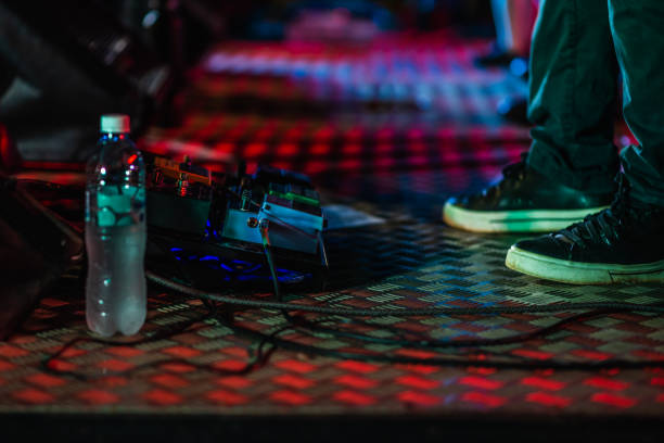 Close-up of a musician's feet wearing black sneakers standing on a textured metal stage floor. A pedalboard with multiple guitar effects pedals and a plastic water bottle are visible. Stage lights cast red and blue reflections