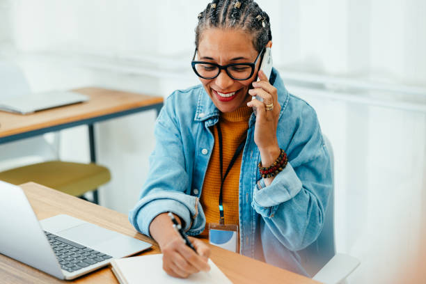 Successful female entrepreneur on a phone call in her modern office, taking notes with a smile