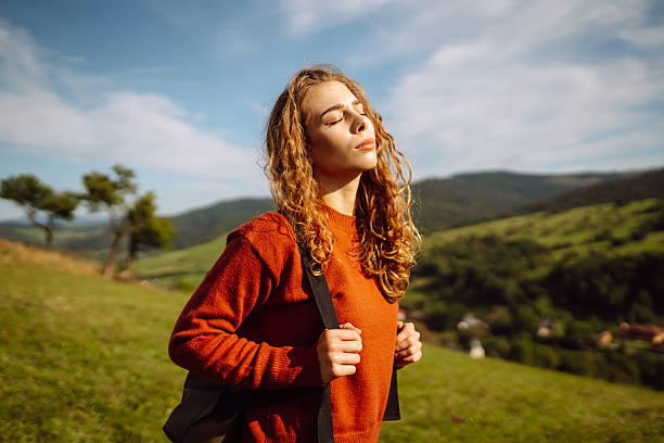 A young woman with a backpack is walking on a green hill. A female traveler in a hat is enjoying the sunny weather, feeling free. Adventure concept, freedom. Active lifestyle.
