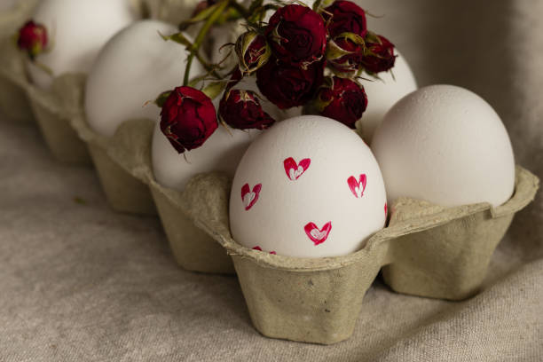 istock White chicken eggs with painted pink hearts in cardboard box, dry red rose 2203641129