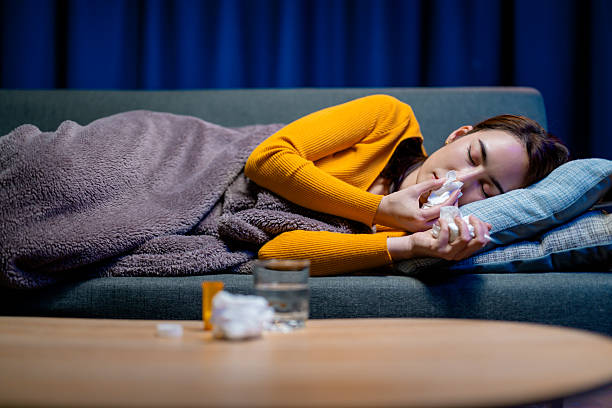 istock Exhausted woman resting on sofa, wrapped in blanket, blowing nose with tissue, surrounded by cold medicine and water glass during nighttime illness 2203645209