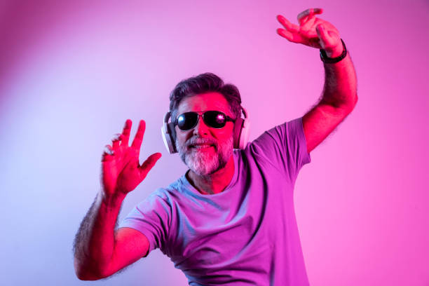 Enjoying his favorite music. Enjoying his favorite music. Happy mature stylish man sunglasses with headphones listening and smiling while standing against pink, blue neon background gay man men sunglasses party stock pictures, royalty-free photos & images